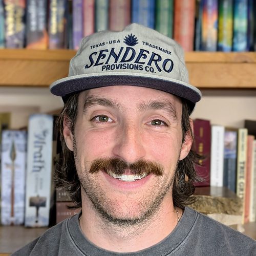 Headshot of Will Luft, a smiling man with a mustache and a ball cap in front of a bookshelf